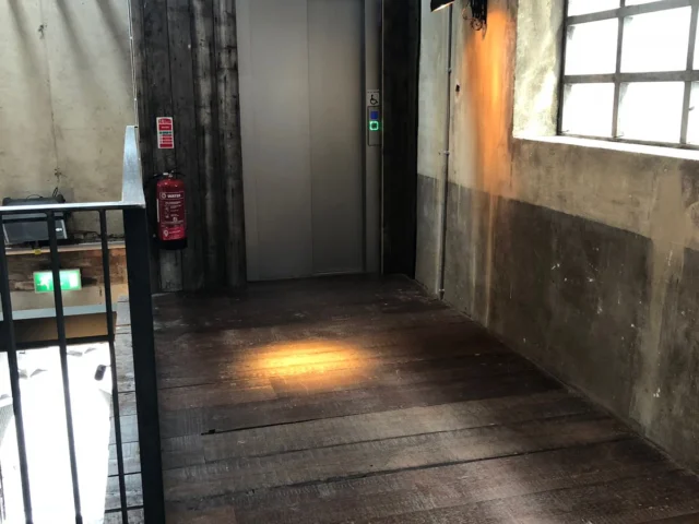 A rustic hallway with wooden floors and exposed beams leads to a modern elevator door. Sunlight streams through a window, casting light spots on the floor.