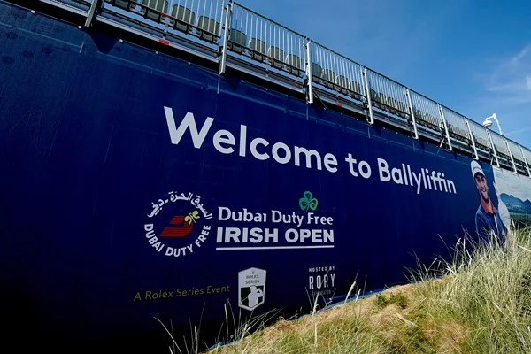 Large sign reading "Welcome to Ballyliffin" with Dubai Duty Free Irish Open branding, event logos, and a partial image of a golfer, situated next to a structure with railings above.
