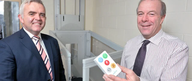 Two men in business attire stand indoors; one holds a control panel with colored buttons, and a lift mechanism is visible in the background.