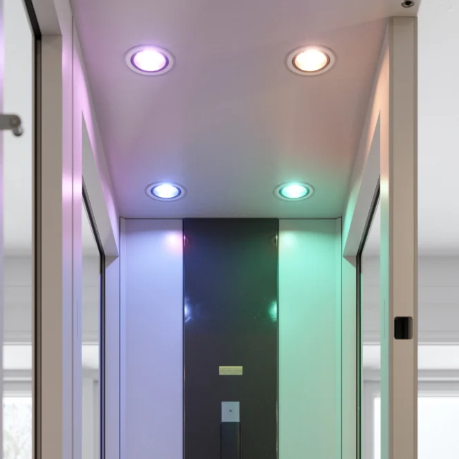 Een smalle gang met vier plafondlampen, die elk een andere kleur uitstralen: paars, oranje, blauw en groen. De muren en het plafond zijn wit en hebben reflecterende oppervlakken.