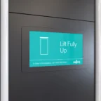 Op een digitaal bord aan de muur staat een liftsymbool met de tekst „Lift Fully Up“ en daaronder een noodnummer.