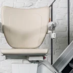A beige stairlift chair is attached to a rail on a staircase next to a white brick wall.