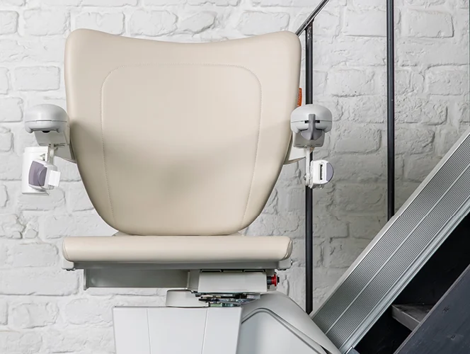 A beige stairlift chair is attached to a rail on a staircase next to a white brick wall.