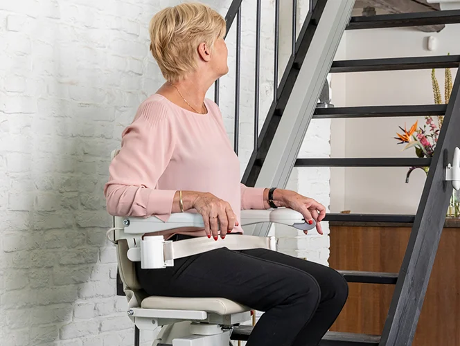 An older woman sits on a stairlift at the base of a staircase, looking to her left in a modern home interior.