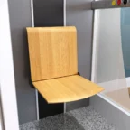 Aan een muur naast een glazen paneel in een lift met een grijs gespikkelde vloer is een opvouwbare houten stoel bevestigd.
