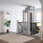 Ein moderner Personenaufzug mit Glaswänden ist neben einer Treppe in einem hellen, weiß getäfelten Flur mit dunklen Geländern und moderner Einrichtung installiert.