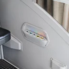 Gros plan sur un panneau de commande comportant six boutons lumineux, fixé sur un mur gris à côté d'un siège noir et d'un rideau, dans un intérieur moderne.