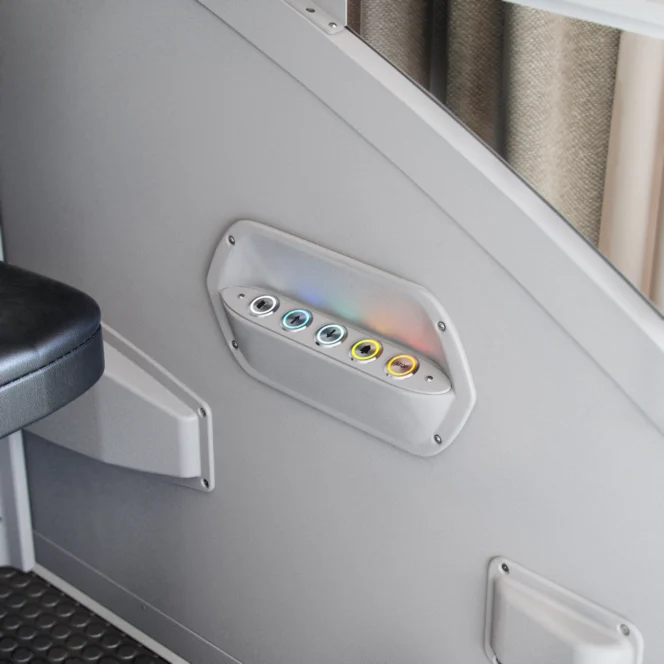 A control panel with six illuminated buttons in different colors is mounted on a gray wall next to a black seat.