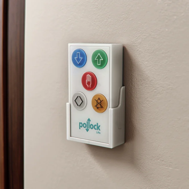 A wall-mounted Pollock Lifts remote control with colored buttons for up, down, stop, door open/close, and alarm functions.