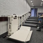 A motorized platform lift is installed on a wall next to a ramp and stairs in an indoor hallway, providing accessibility for wheelchairs.