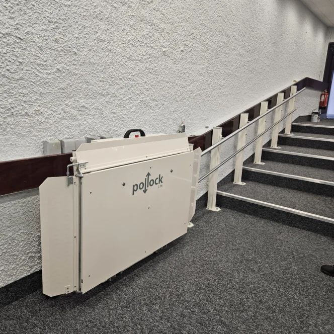 A folded Pollock platform lift is mounted beside a set of carpeted stairs with a handrail in a hallway.