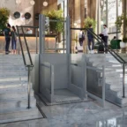 A modern hotel lobby with a platform wheelchair lift installed between two sets of marble stairs, with people conversing and checking in at the reception desk.