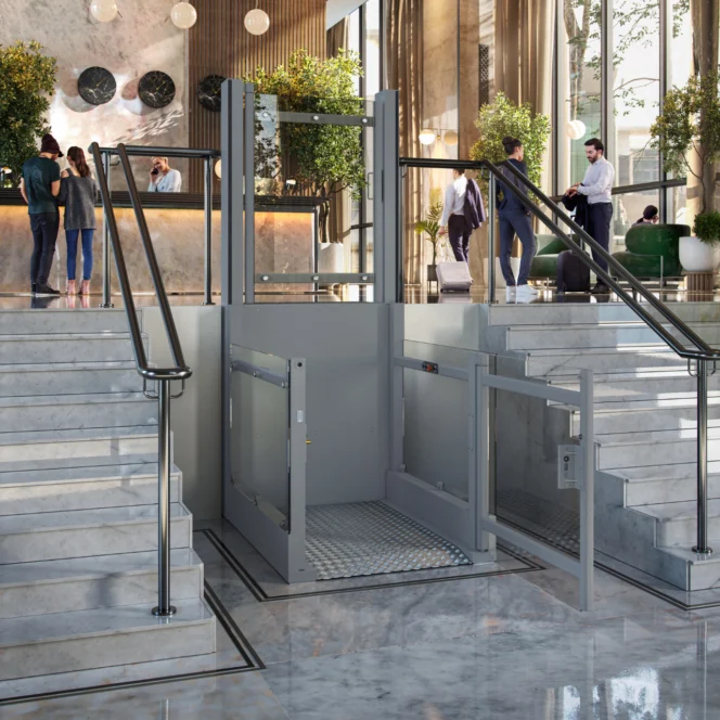 A modern hotel lobby with a platform wheelchair lift installed between two sets of marble stairs, with people conversing and checking in at the reception desk.