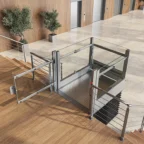 A modern indoor wheelchair platform lift with glass panels is installed beside a staircase in a building lobby with elevators and potted plants.