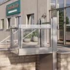A platform lift with glass panels installed outside a building with an SJ Bargh LTD sign, providing accessible entry from ground level.