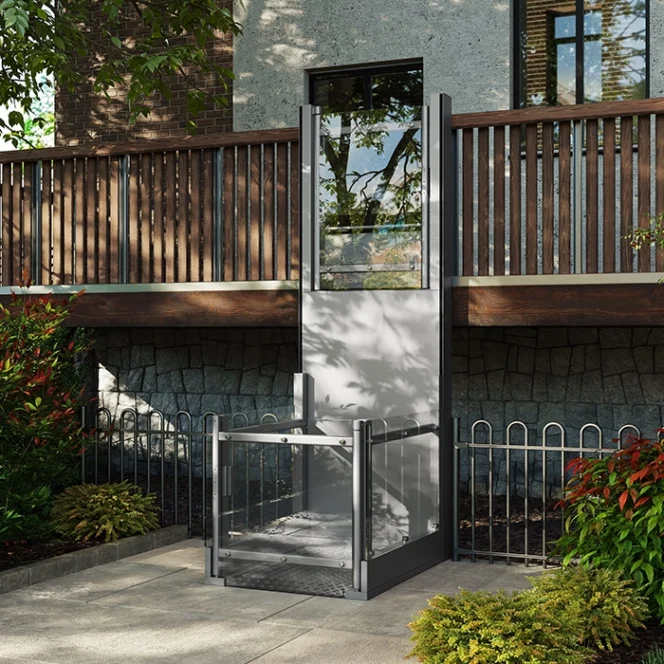 A vertical platform lift is installed outdoors by a wooden deck, providing accessible entry to an elevated area. Plants and a stone wall are visible nearby.