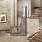 A modern residential lift is installed beside a staircase in a spacious, well-lit foyer with beige stone walls and elegant decor.