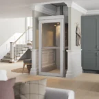 Un ascenseur domestique moderne doté de portes vitrées est installé dans un espace de vie lumineux et ouvert, avec un parquet en bois clair et des murs blancs.