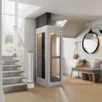 A modern home elevator installed beside a staircase in a bright hallway with light wood floors, a bench, a round mirror, and wall hooks.
