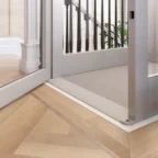 Ein moderner Hausaufzug mit Türen aus Glas und Metall führt auf einen hellen Holzboden im Fischgrätenmuster in der Nähe einer Treppe mit dunklen Geländerstäben.