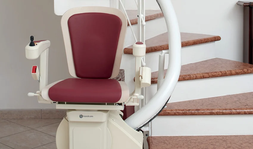 A motorized stair lift with a maroon seat and control panel is installed beside a curved staircase with brown steps.