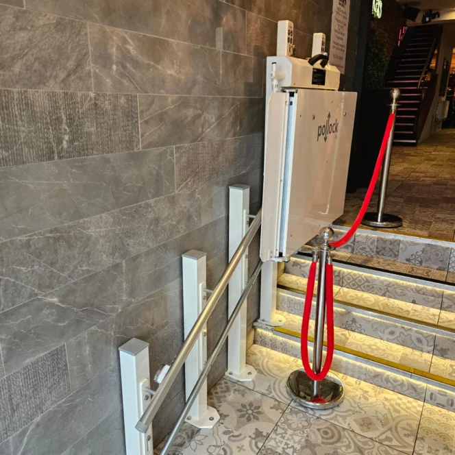 A platform lift for wheelchair access is installed beside a small staircase with a red velvet rope barrier in a tiled interior space.