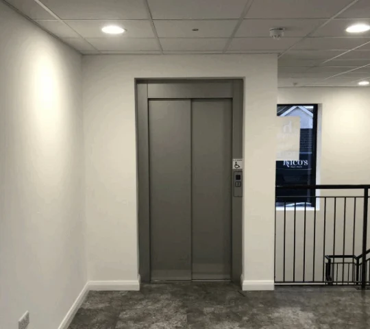 A closed elevator with metal doors is centered in a hallway with white walls, gray flooring, and a nearby stair railing on the right.