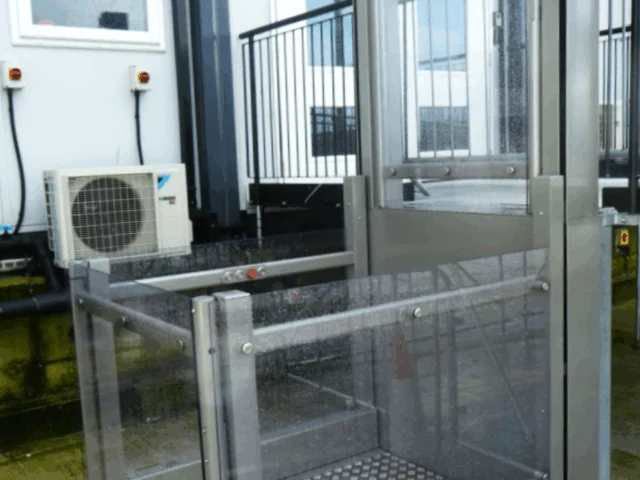 A metal and glass outdoor platform lift is installed next to a building with steps, providing wheelchair accessibility.