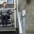 A woman in a wheelchair uses an outdoor stairlift platform to move up a set of concrete stairs next to a rough-textured wall.