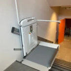 A stair lift platform for wheelchair users is installed on a staircase next to a wall, with an orange door and a chair visible in the background.