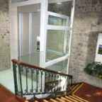 Glass elevator with metal doors and control panel situated next to a staircase with a black railing, in a stone-walled interior.