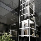 A man stands inside a modern glass elevator within a contemporary building featuring office and closet spaces.