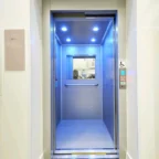 An open modern elevator with blue ceiling lights, metallic handrails, and accessible buttons, viewed from a building corridor.