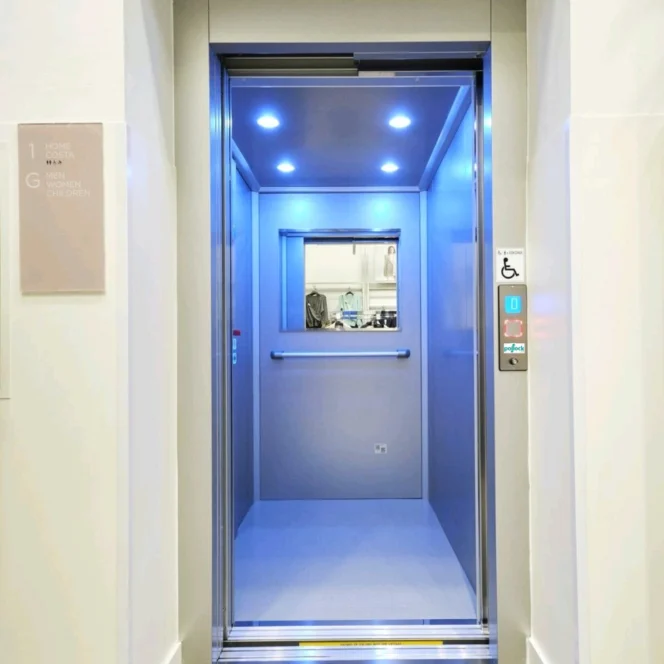 An open modern elevator with blue ceiling lights, metallic handrails, and accessible buttons, viewed from a building corridor.