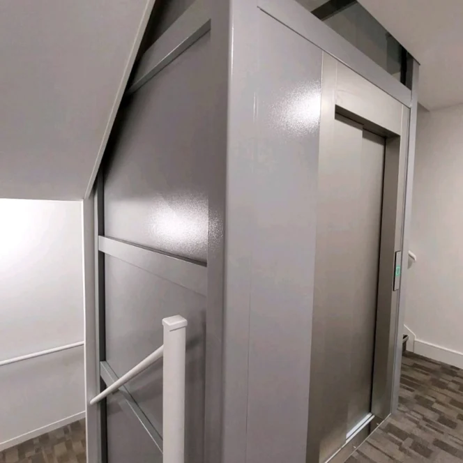 A modern elevator is installed beside a staircase in a building corridor, with gray paneling and glass panels above the elevator door.