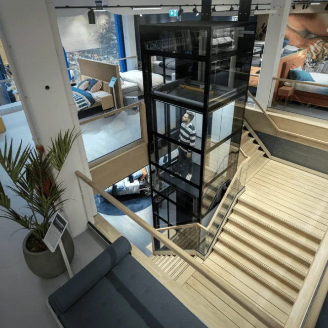 A modern furniture showroom with a central glass elevator, wooden staircase, sofas, display areas, and indoor plants visible.