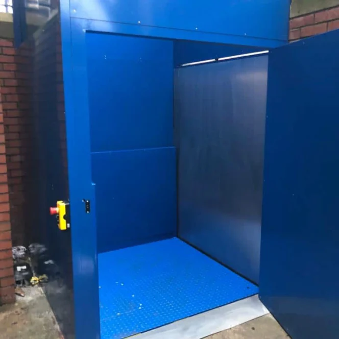 A blue platform lift with open doors, revealing an empty interior and a red emergency stop button on the outside wall.