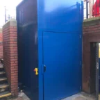 A blue metal enclosure with a door and handle is positioned between brick walls next to a staircase and an outdoor walkway.