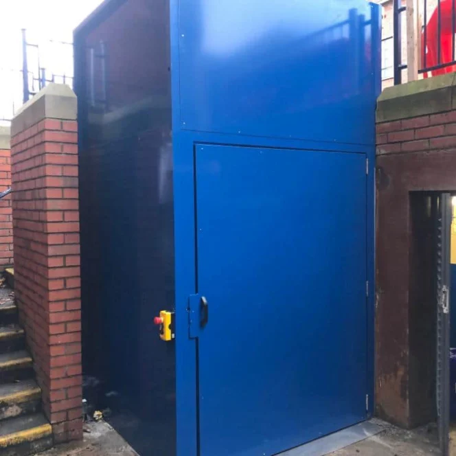 A blue metal enclosure with a door and handle is positioned between brick walls next to a staircase and an outdoor walkway.