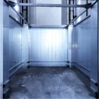 An empty industrial freight elevator with metal walls, ceiling, and floor, illuminated by overhead lighting; control panel visible on the right side.