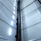 Looking up inside an empty industrial elevator shaft with metal rails and vertical fluorescent lights on the walls.