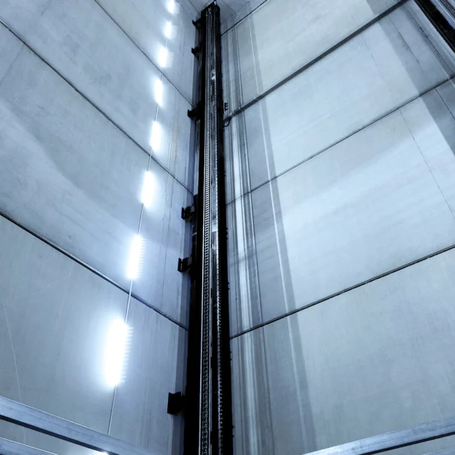 Looking up inside an empty industrial elevator shaft with metal rails and vertical fluorescent lights on the walls.