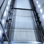 View looking up an empty elevator shaft with visible guide rails, chains, and lights mounted on concrete walls.