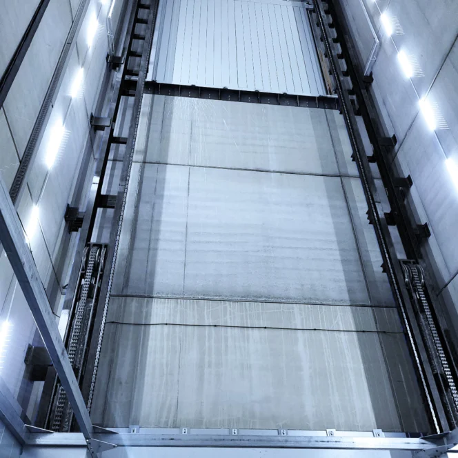 View looking up an empty elevator shaft with visible guide rails, chains, and lights mounted on concrete walls.