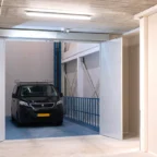 A black van with a yellow license plate is parked inside a modern indoor parking lift with open white doors and concrete ceiling.