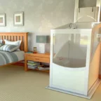 A compact home elevator is installed next to a wooden bed in a tidy bedroom with beige carpeting and light-colored walls.