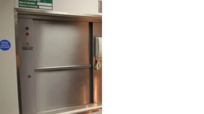 A closed stainless steel serving hatch in a wall, with a green first aid sign and a wall-mounted paper towel dispenser next to it.