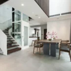 Modern open-plan interior with a staircase, glass elevator, dining table with chairs, potted plant, and minimalistic decor.