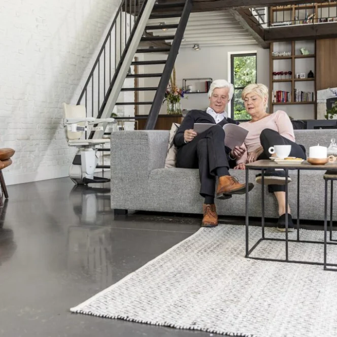 An older couple sits together on a gray sofa in a modern living room, looking at a tablet. Shelves, a staircase, and kitchen area are visible in the background.