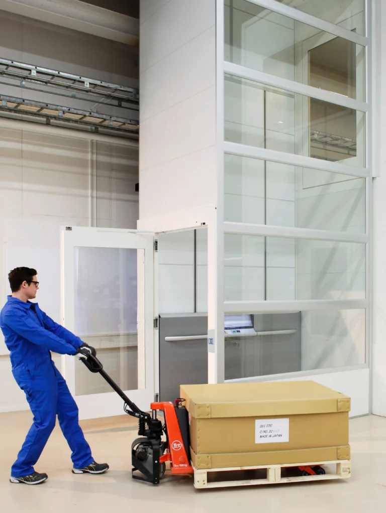 A person in blue coveralls uses a pallet jack to move a large boxed item on a pallet in an industrial indoor setting near a modern elevator.
